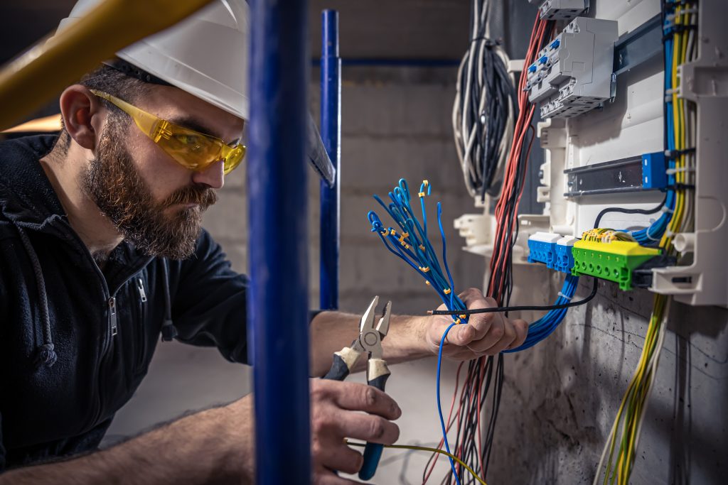 A male electrician works in a switchboard with an electrical connecting cable, connects the equipment with tools.